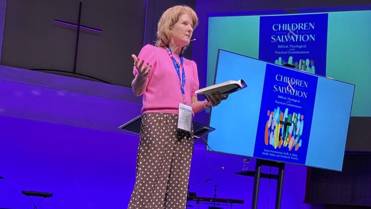 Shelly Melia wearing a pink shirt, standing on a stage speaking